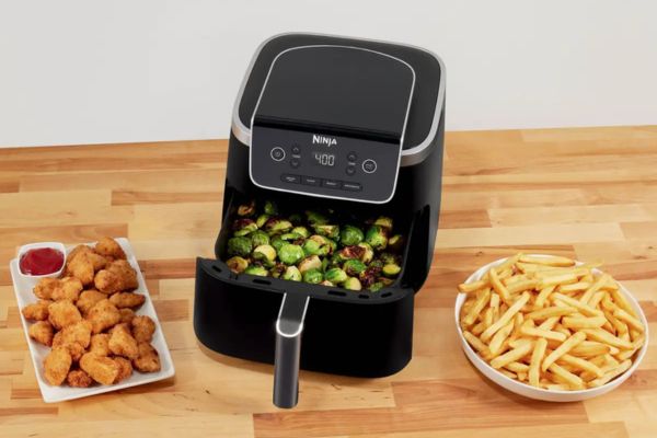 Air frier open with Brussel sprouts inside alongside a plate of French fries and chicken nuggets
